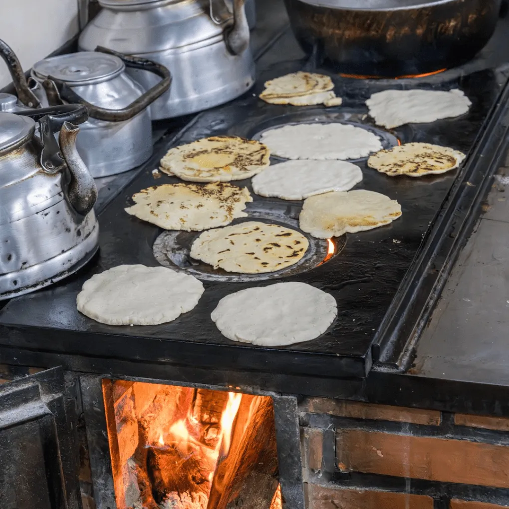 Tortillas cooking on a wood stove, surrounded by kettles and a pot.