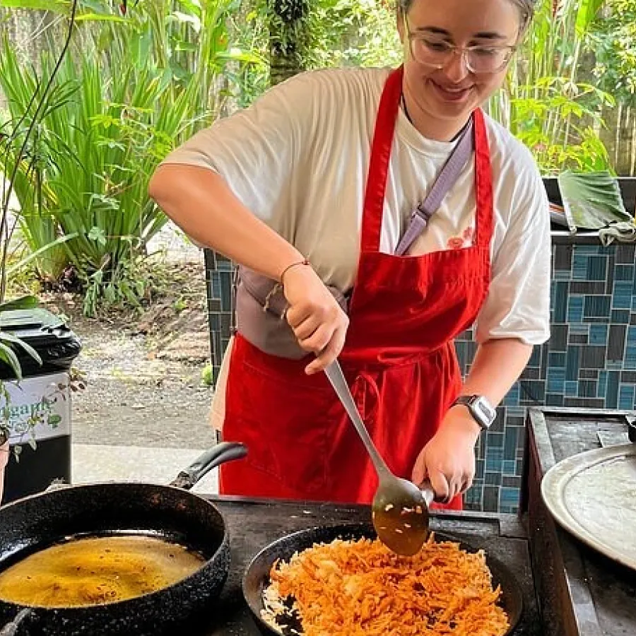 Person cooking rice outdoors, wearing red apron and white shirt, surrounded by plants.