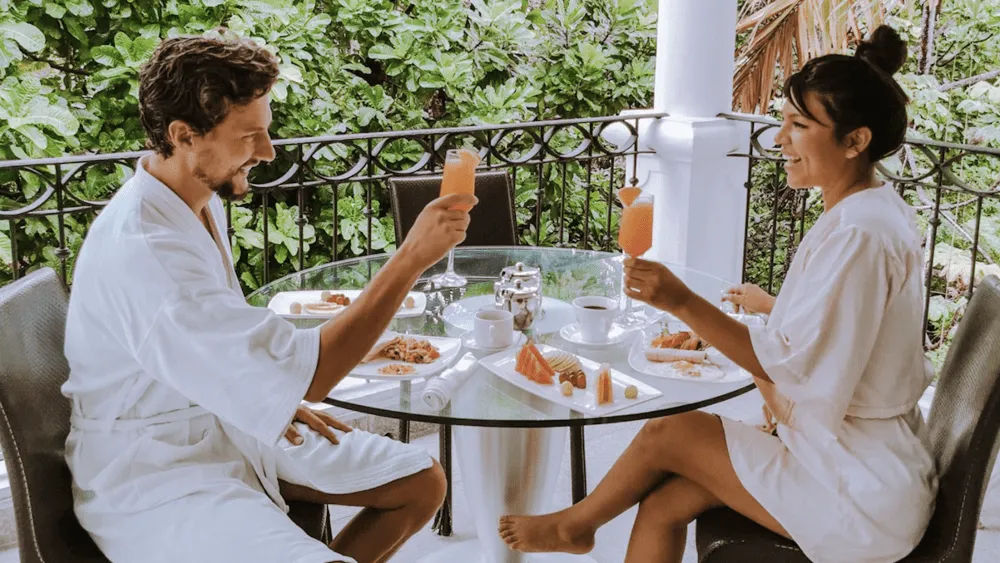 Couple in robes having breakfast on a patio, toasting with orange drinks.