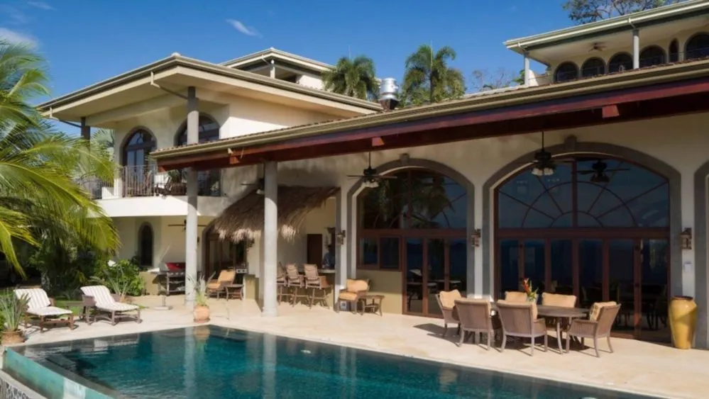 Luxurious villa with infinity pool, patio chairs, and palm trees under a clear blue sky.