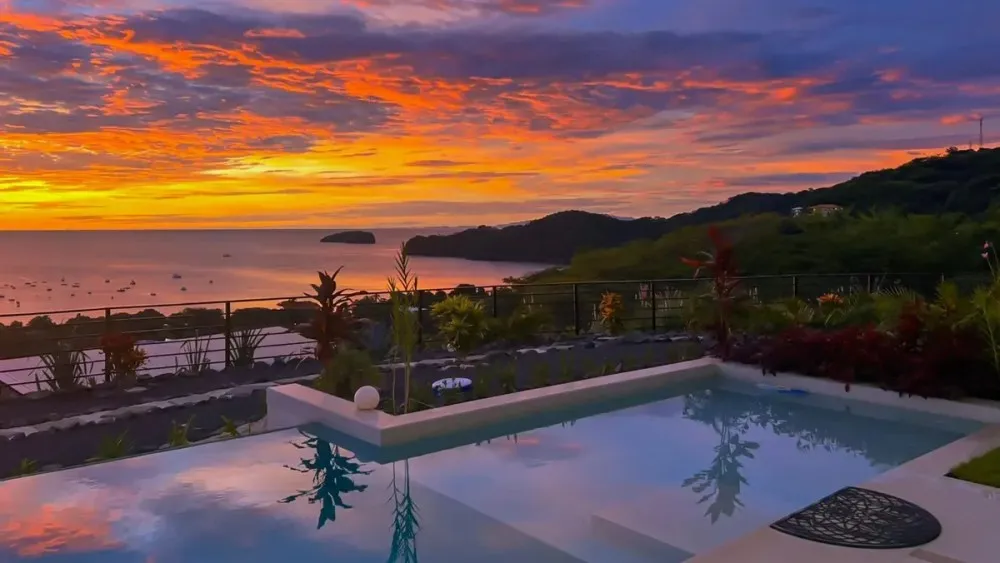 Ocean sunset view with pool and lush hills in the foreground.