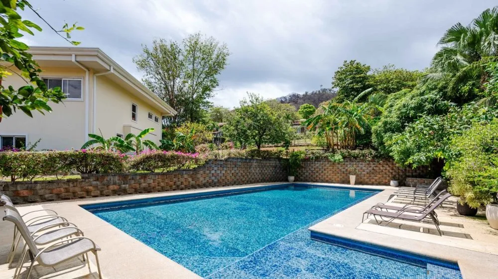 A backyard swimming pool surrounded by chairs and lush greenery.