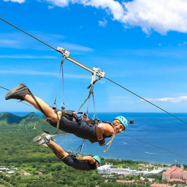 Two people zip-lining over lush landscape with ocean view.
